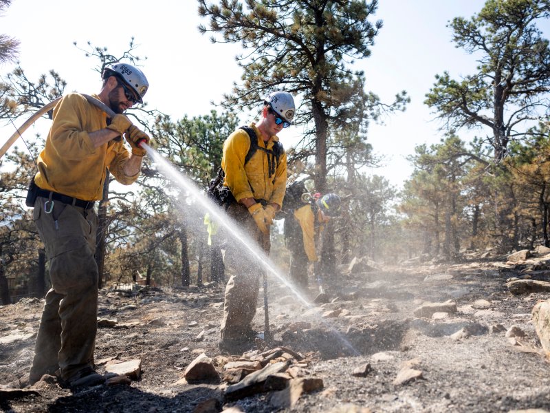 Firefighters in yellow uniforms use a hose to extinguish smoldering ground in a forested area with charred trees and rocks.