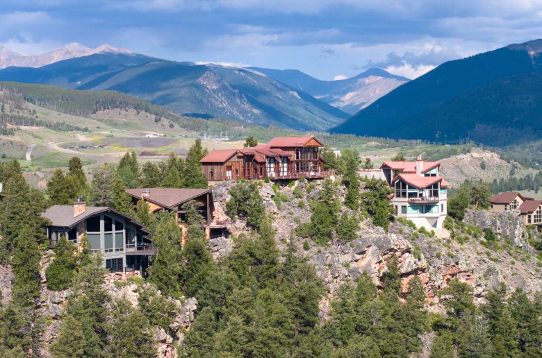 Large houses sit on top a mountain. A mountain range is visible behind them.