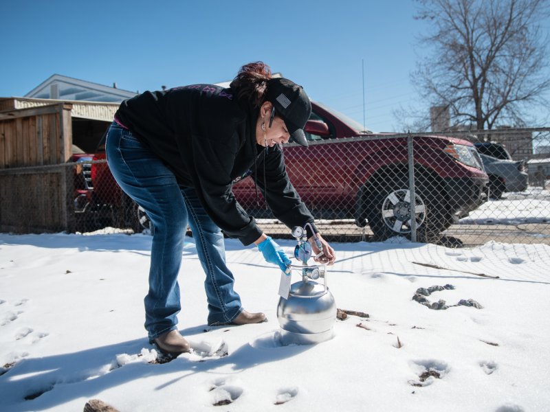 A person in a black jacket and blue jeans collects an air sample using a metal container in a snowy outdoor area near a fenced yard and parked cars.