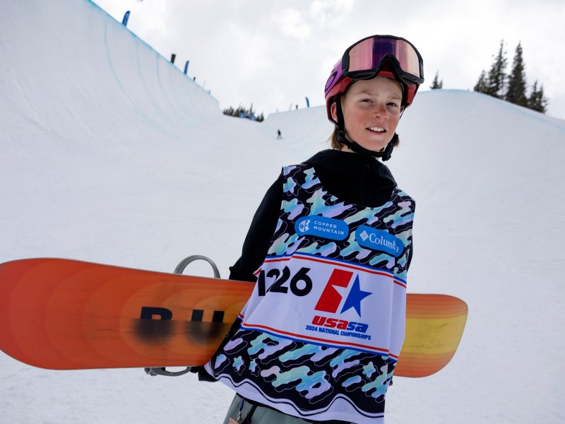 A young boy holds a snowboard outside at the bottom of a halfpipe