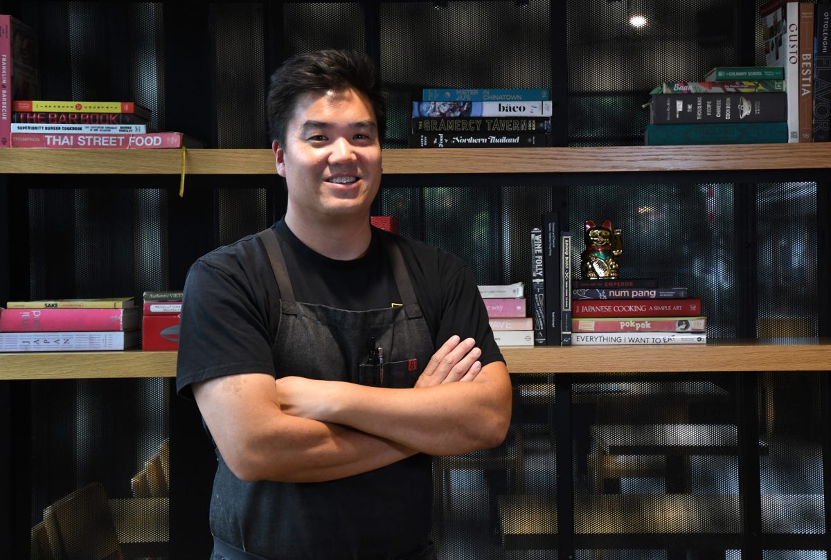 Tommy Lee with crossed arms stands in front of a bookshelf filled with books and a small golden cat figurine.