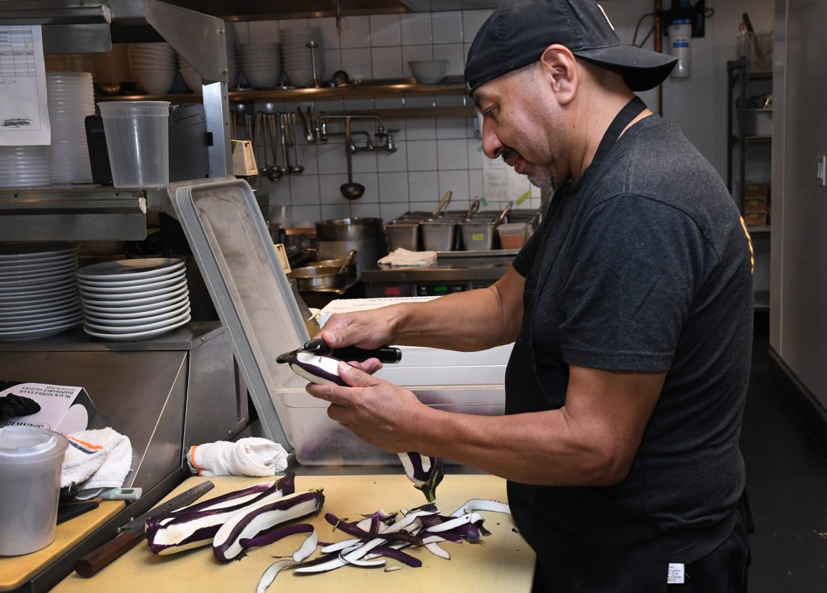 A man in a black cap and apron peels an eggplant in a stainless steel kitchen with various cooking utensils and dishes in the background.