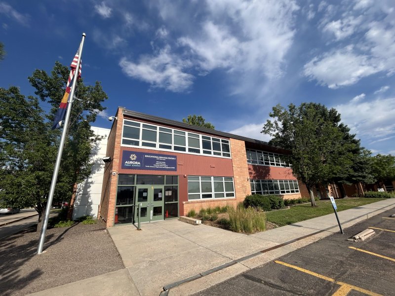 A two-story brick building with large windows and a sign reading "Aurora". An American flag is hoisted on a flagpole in the foreground. Bright blue sky with scattered clouds overhead.