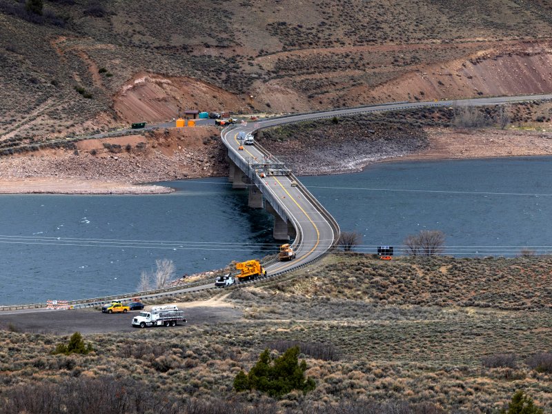 The U.S. 50 bridge under construction spans Blue Mesa Reservoir. Construction vehicles and equipment are on and near the bridge with machinery and traffic cones indicating ongoing work. Bushes and hills surround the area.