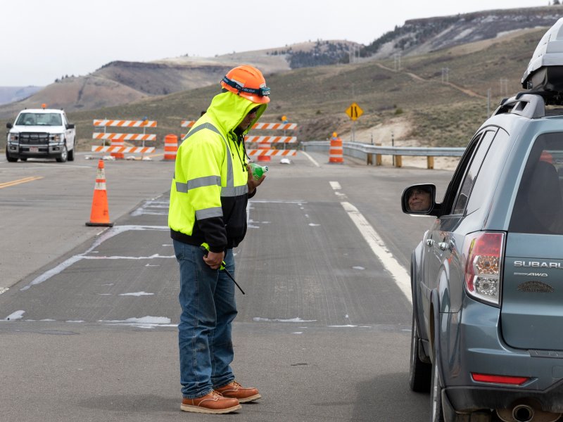 a man in neo green jacket and jeans with orange helmet stands next to a car on a highway