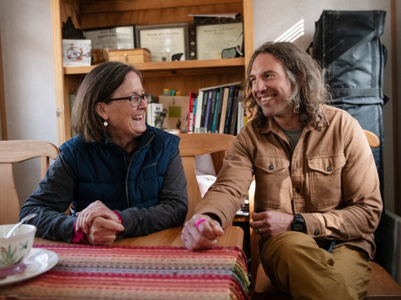 Jill Dreves, wearing a puffer vest over a long sleeved shirt, leans on a table while looking at and laughing with Justin Gold, who is sitting next to her in a tan longsleeved shirt.