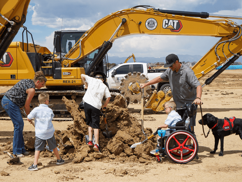 People, including a child in a wheelchair, participate in a groundbreaking event for inclusive parks, with construction equipment in the background. A service dog stands beside the child in the wheelchair.