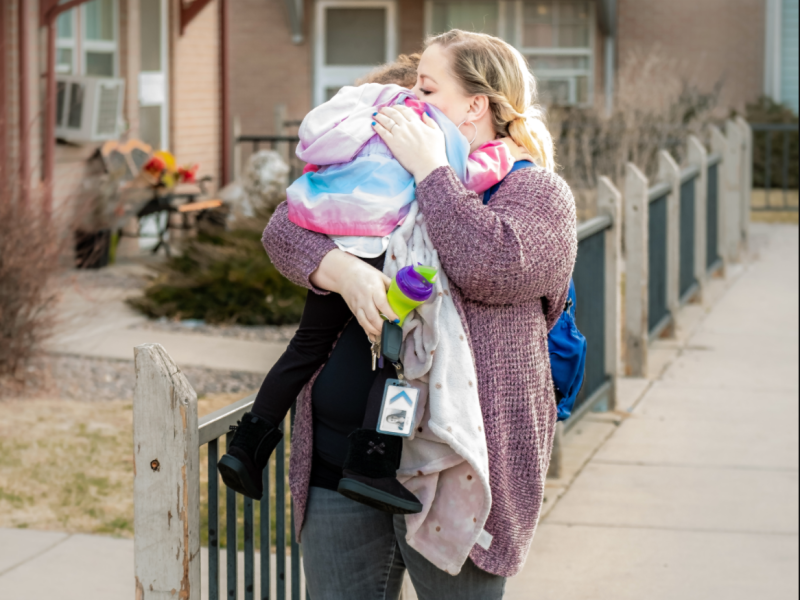 A woman holding a child in her arms kisses the child's head while standing outside near a fence and building. The woman is carrying a water bottle and a blue backpack.