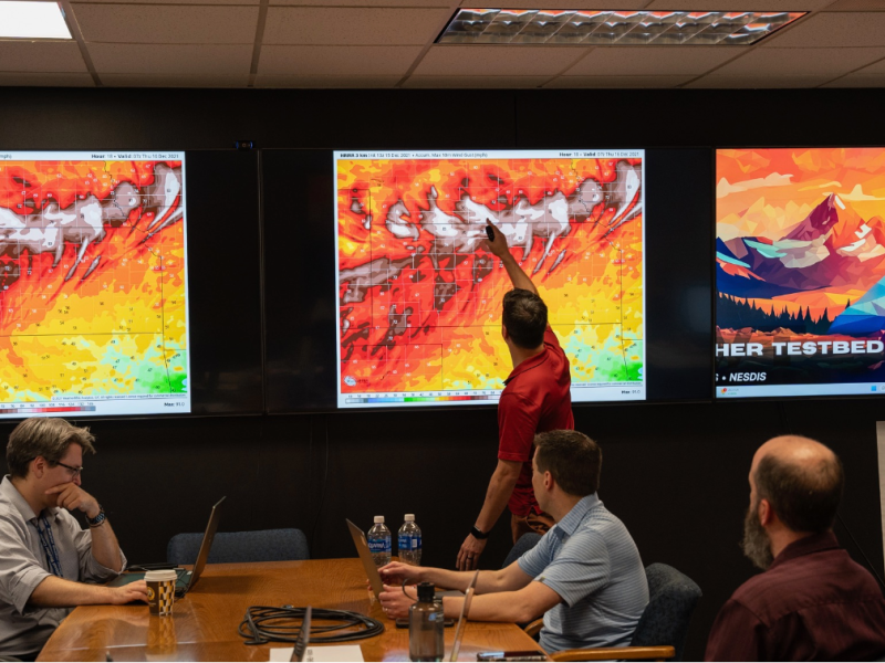 A group of four people in an office room observes weather maps displayed on three large screens, with one person pointing to a screen. A whiteboard with writing is visible on the right side.