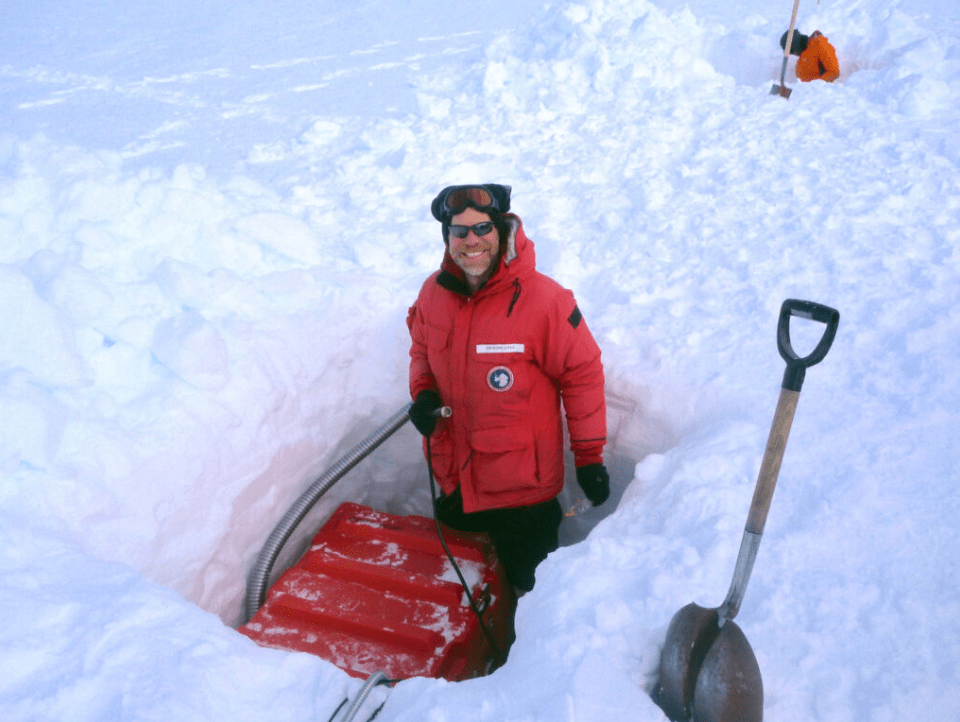 Rick Aster, in red winter gear, standing in a snow trench with equipment, smiling. Shovel is stuck upright in the snow beside him.