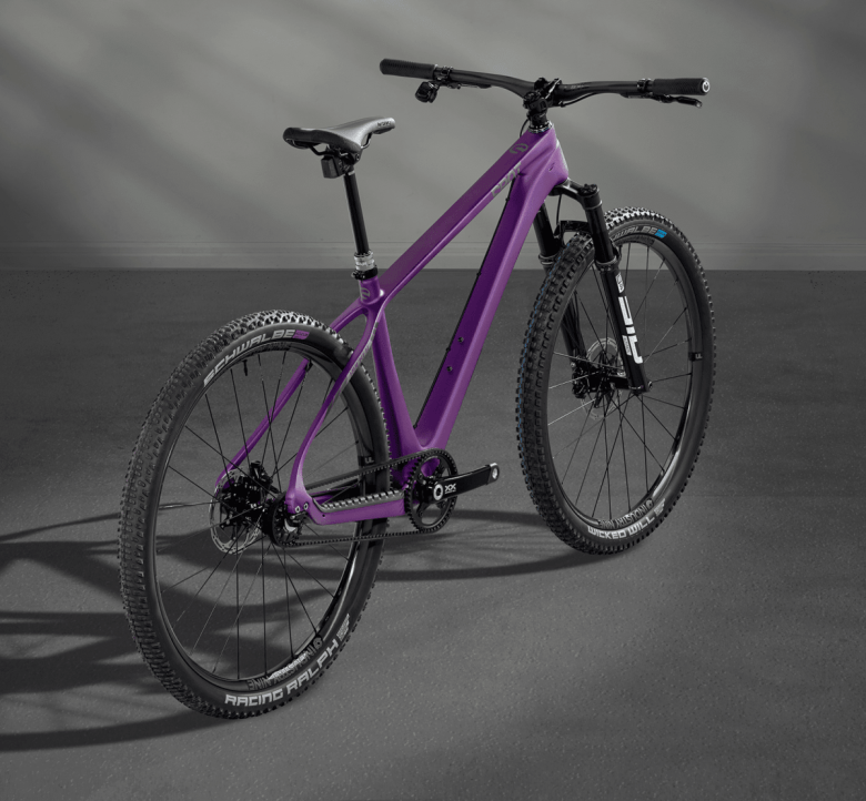 A purple mountain bike with knobby tires, a front suspension fork, and a black seat stands on a flat gray surface. Instead of a typical bike chain, the bike has a belt.