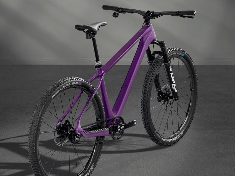 A purple mountain bike with knobby tires, a front suspension fork, and a black seat stands on a flat gray surface. Instead of a typical bike chain, the bike has a belt.